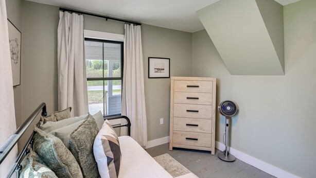 A bedroom with a bed, a dresser, a standing fan, and a window with curtains.