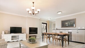 A living room with a TV, a fireplace, a dining area with chairs, and a kitchen with cabinets. - Winelands Golf Lodges 10 (Cape Town)