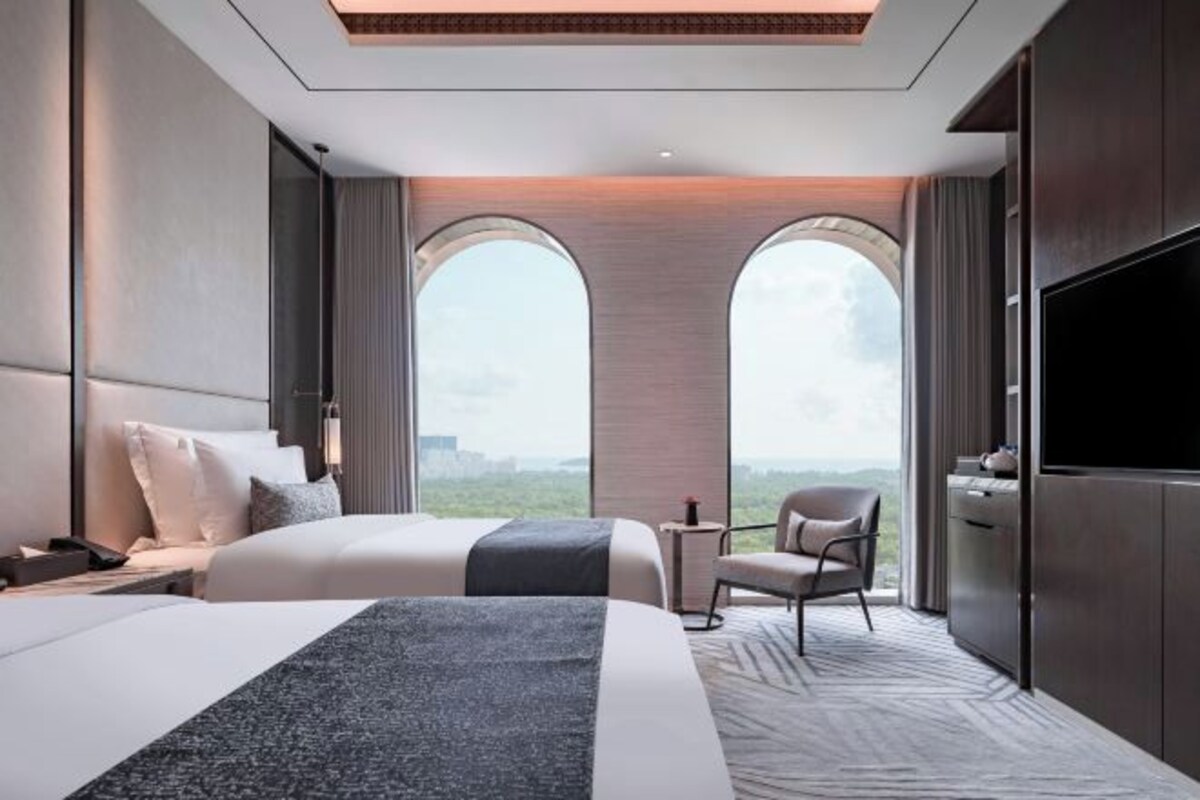 A modern hotel room with a large bed, a desk, and a TV. There are two large windows with a view of the city.