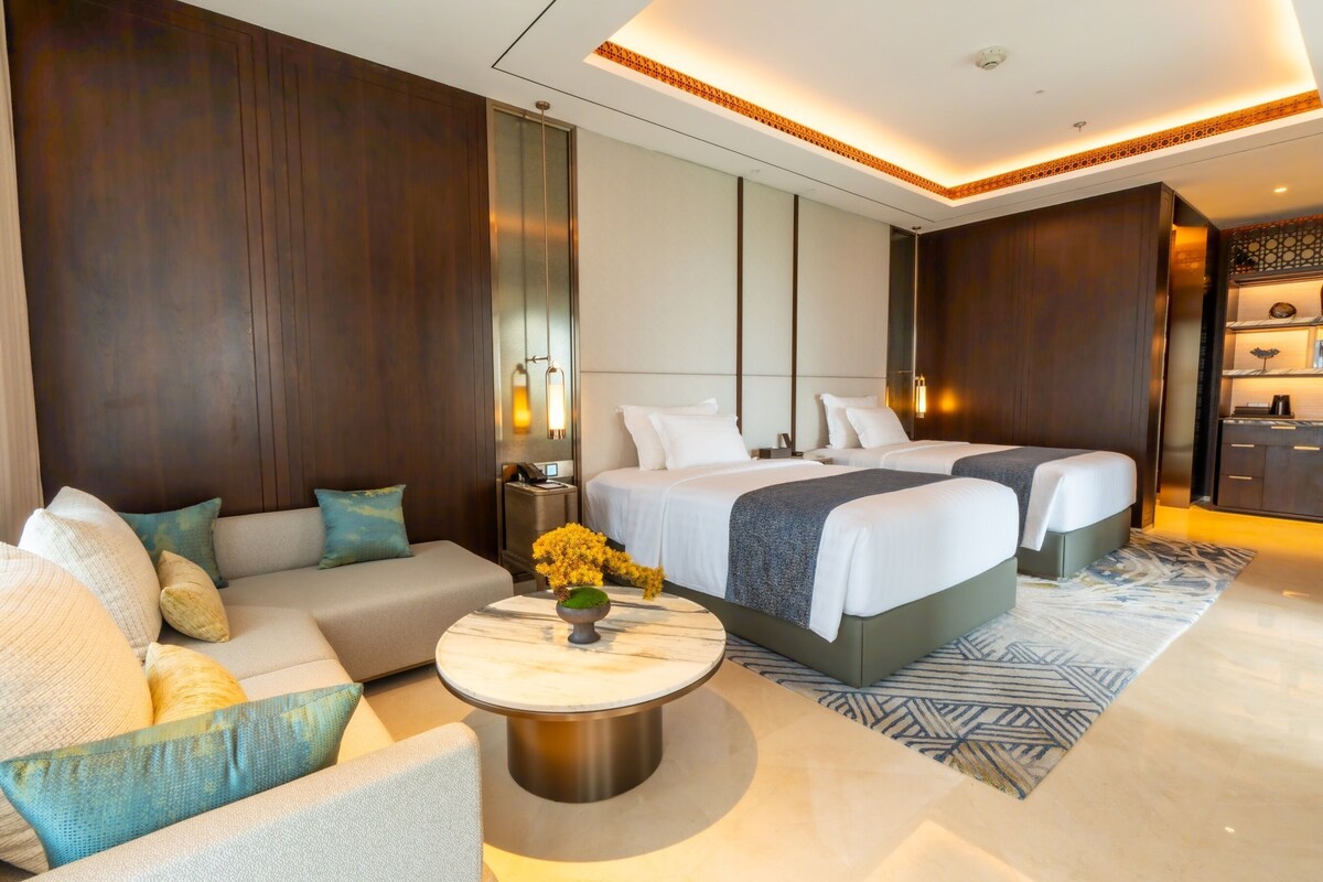 A modern hotel room with a sofa, two beds, a nightstand, and a decorative vase.