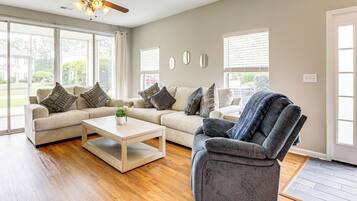 A living room with two sofas, a coffee table, and a recliner. There are large windows with blinds, a ceiling fan, and a door to the right.