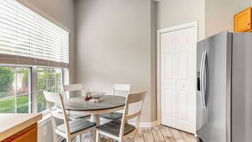 A kitchen with a round table, white chairs, a stainless steel refrigerator, and a window with blinds.