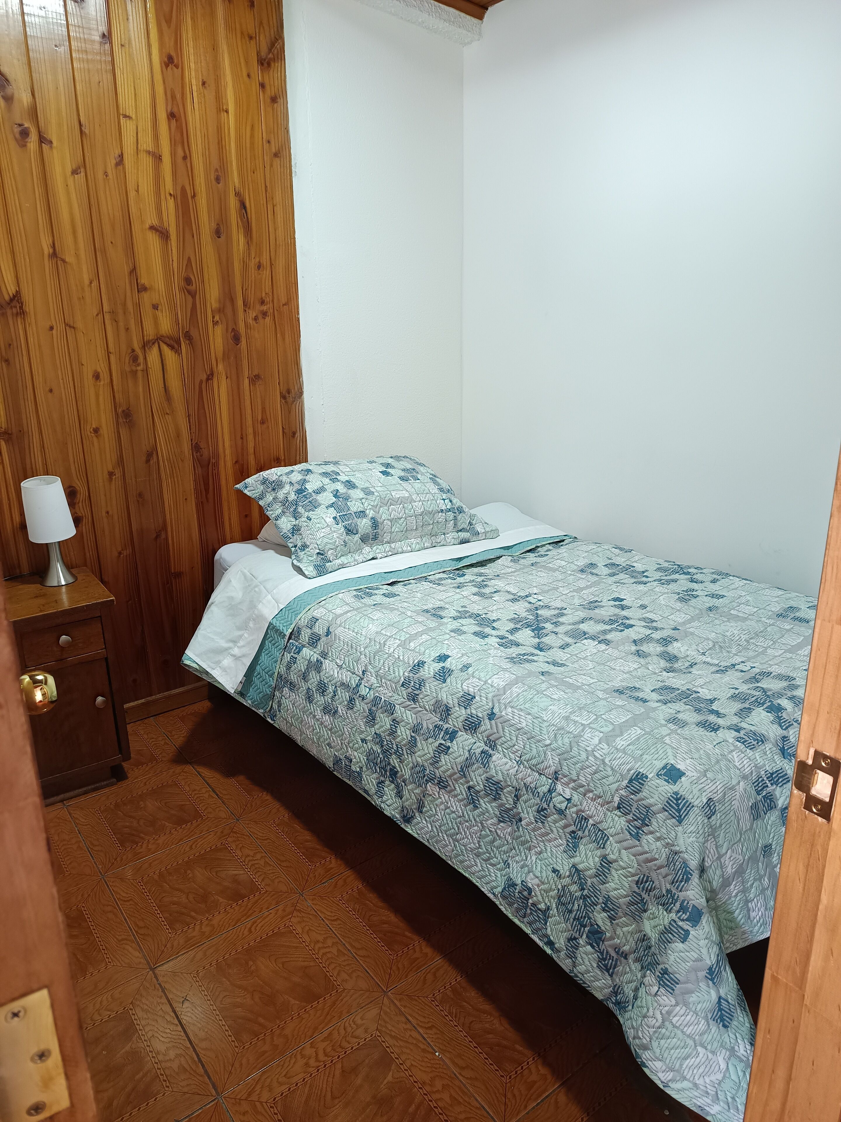Ein kleines, ordentlich eingerichtetes Schlafzimmer mit Holzboden, einem Einzelbett mit gemusterten Betttextilien, einem Nachttisch mit Lampe und einem Holzschrank.