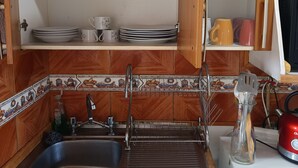 Una cocina con armarios de madera, un lavamanos y una estufa. Hay repisas sobre el lavamanos con vajilla y un saco de azúcar.