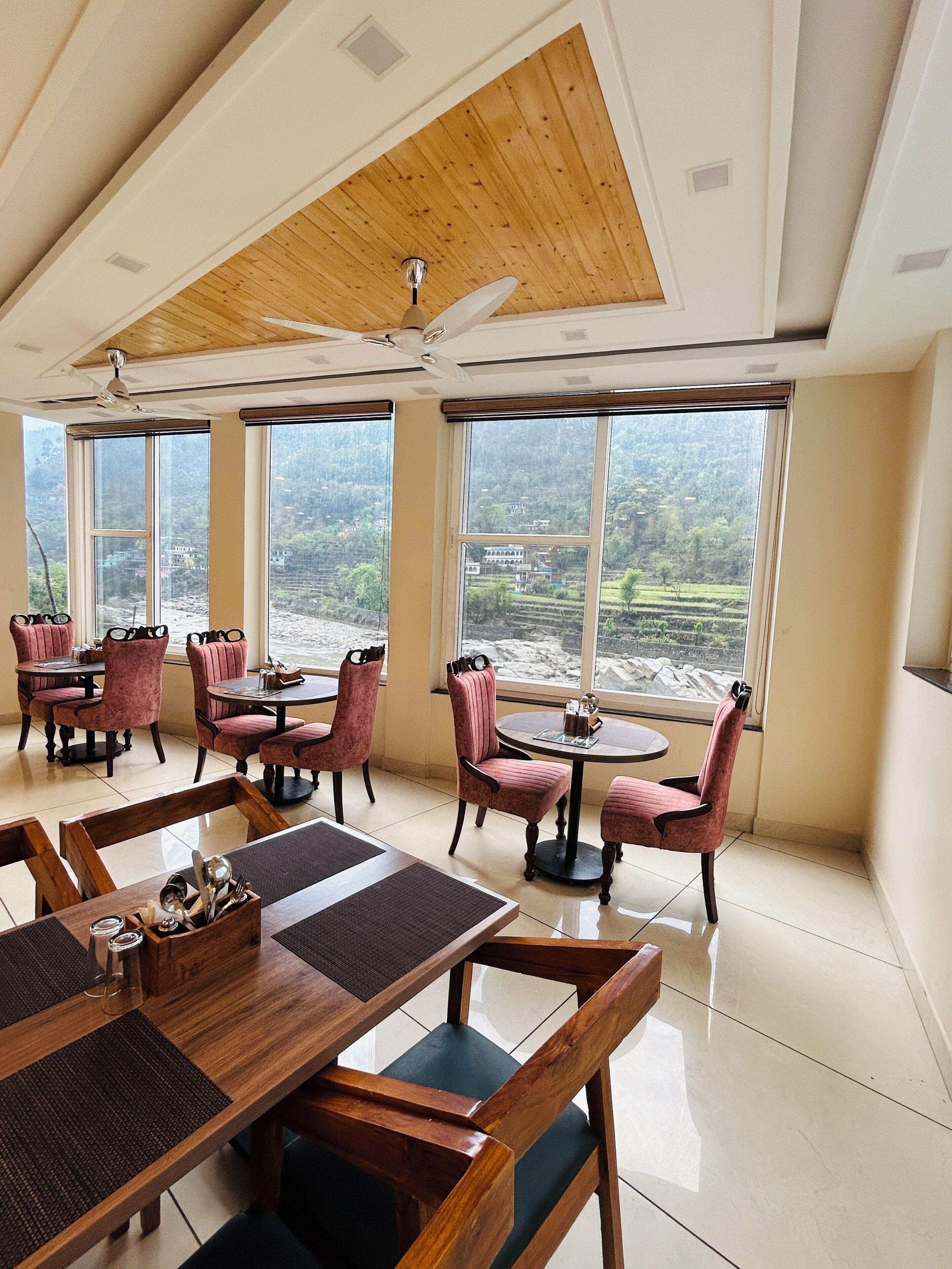 Un comedor con mesas y sillas de madera, ventanas amplias con vistas a un paisaje montañoso y un ventilador de techo.