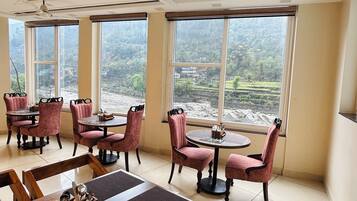Un comedor con mesas y sillas de madera, ventanas amplias con vistas a un paisaje montañoso y un ventilador de techo.