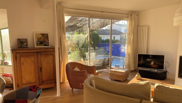 A living room with a sofa, a television, a wicker chair, a cabinet, and a view of a swimming pool through large windows. - House near Montpellier quiet and bright swimming pool (Laverune)