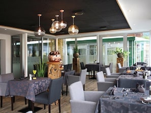 A restaurant with multiple tables and chairs, large windows, and hanging pendant lights. - Gutjahr (Abtenau)
