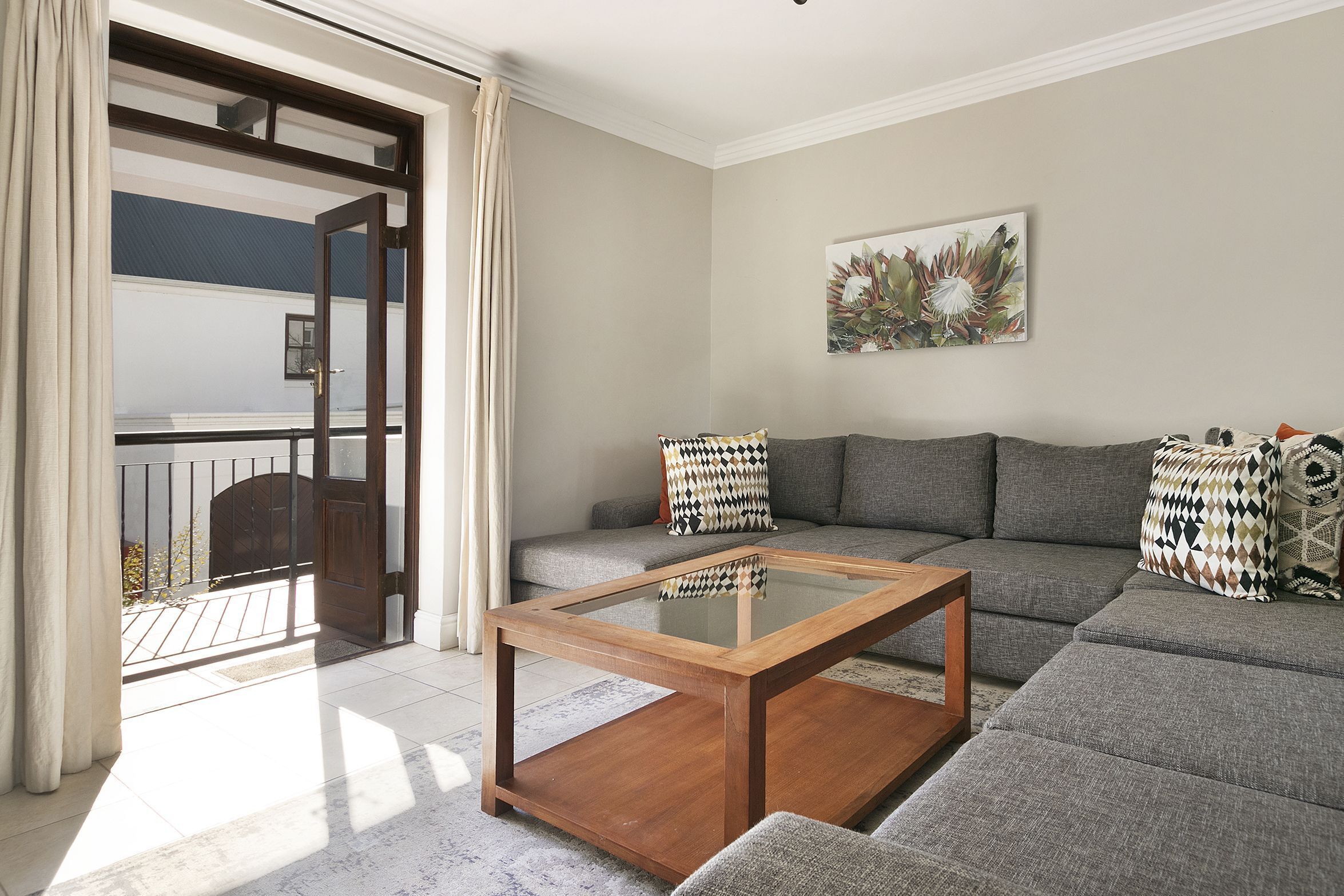 Una sala de estar moderna con una mesa de centro de vidrio, sofá gris, cojines estampados y un ventanal con cortinas.