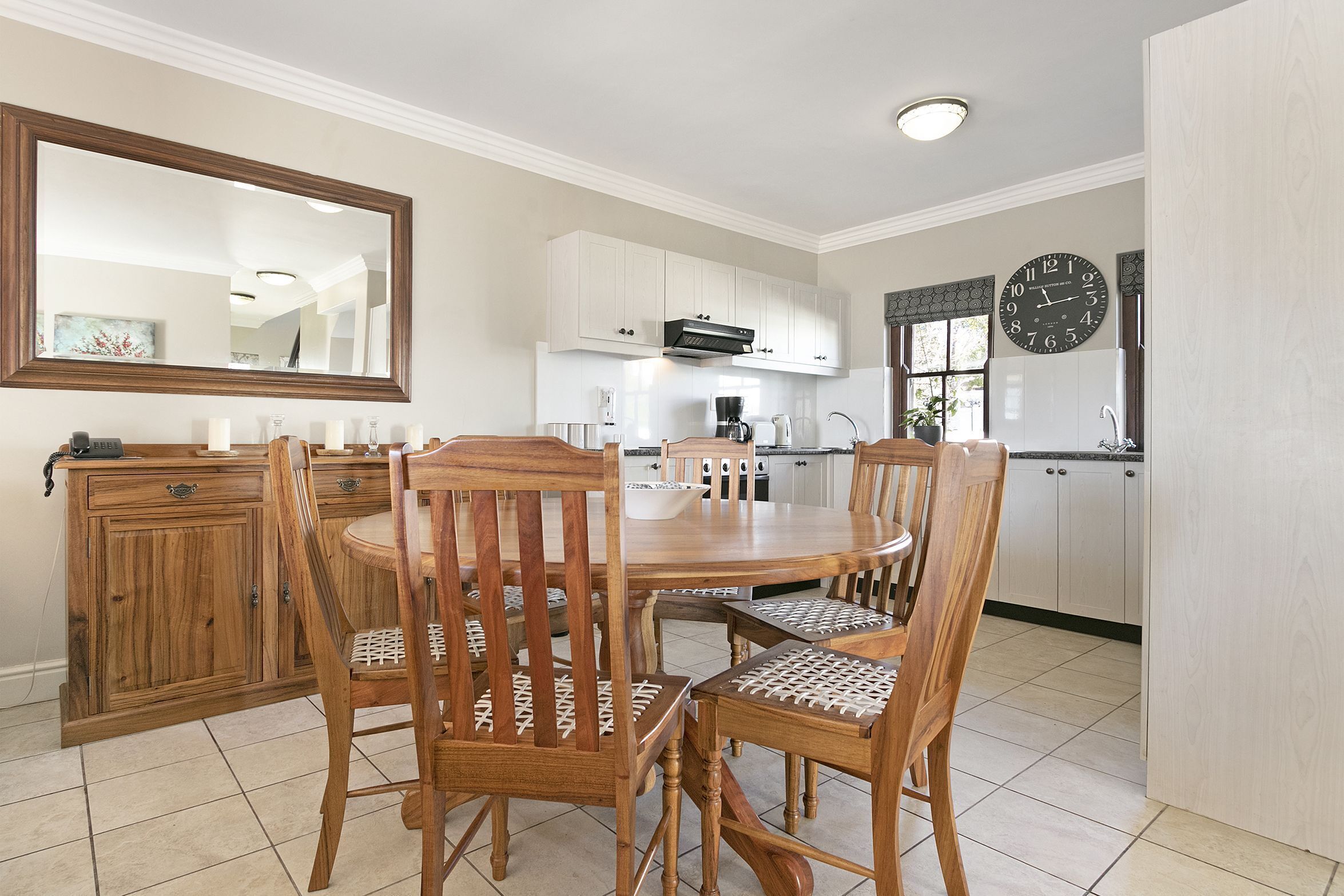Un coin repas avec une table ronde et quatre chaises en bois, un buffet en bois, un grand miroir et, au fond, une cuisine.