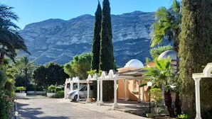 Ein Kiesweg führt zu einem Gebäude mit weißen Säulen und einer Kuppel, umgeben von Grünpflanzen und Bergen im Hintergrund.