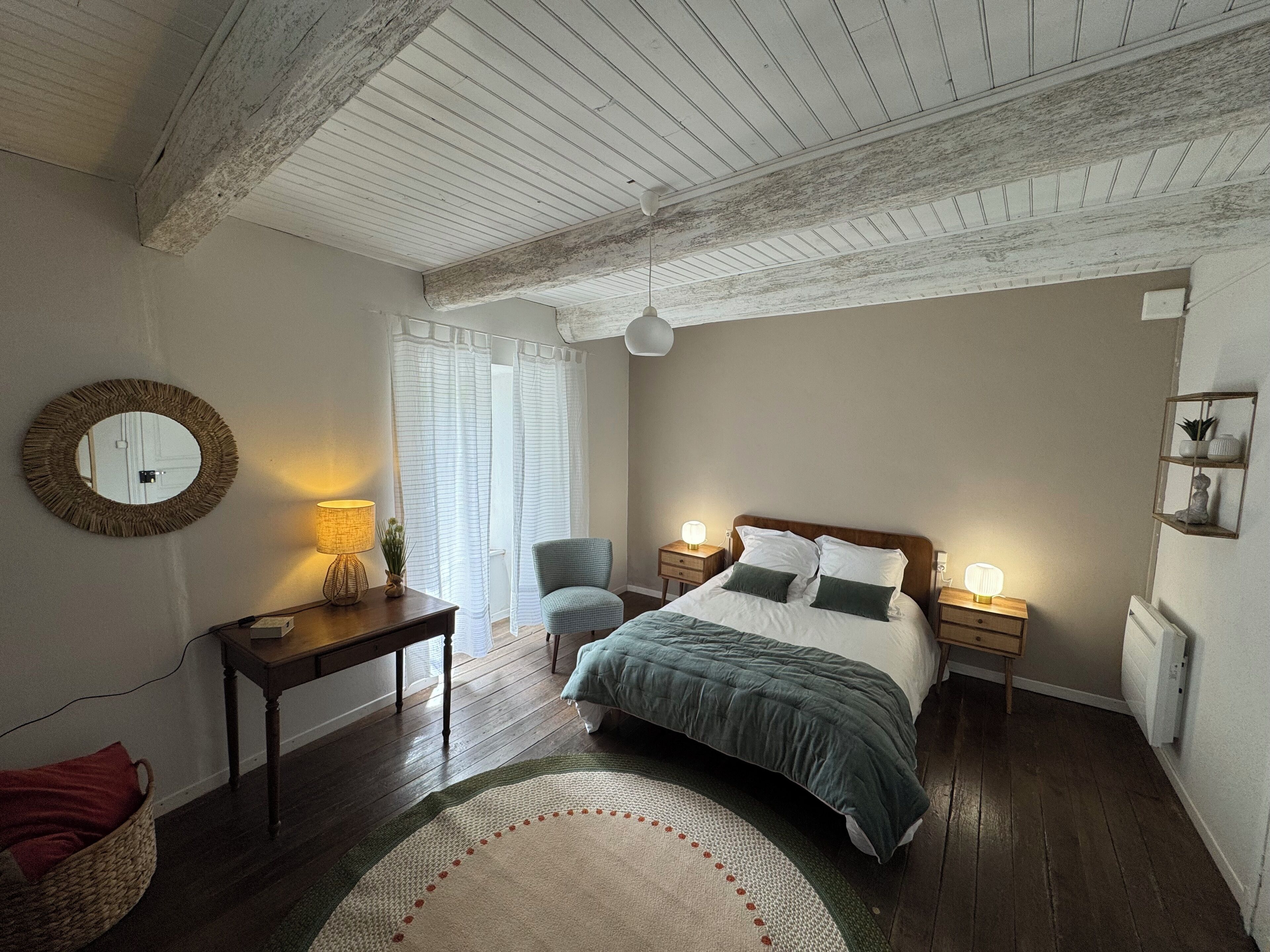 Une chambre avec un plafond en bois, un lit recouvert d’une couette verte et blanche, deux lampes de chevet, un miroir rond, une petite table avec une lampe et un panier en osier.