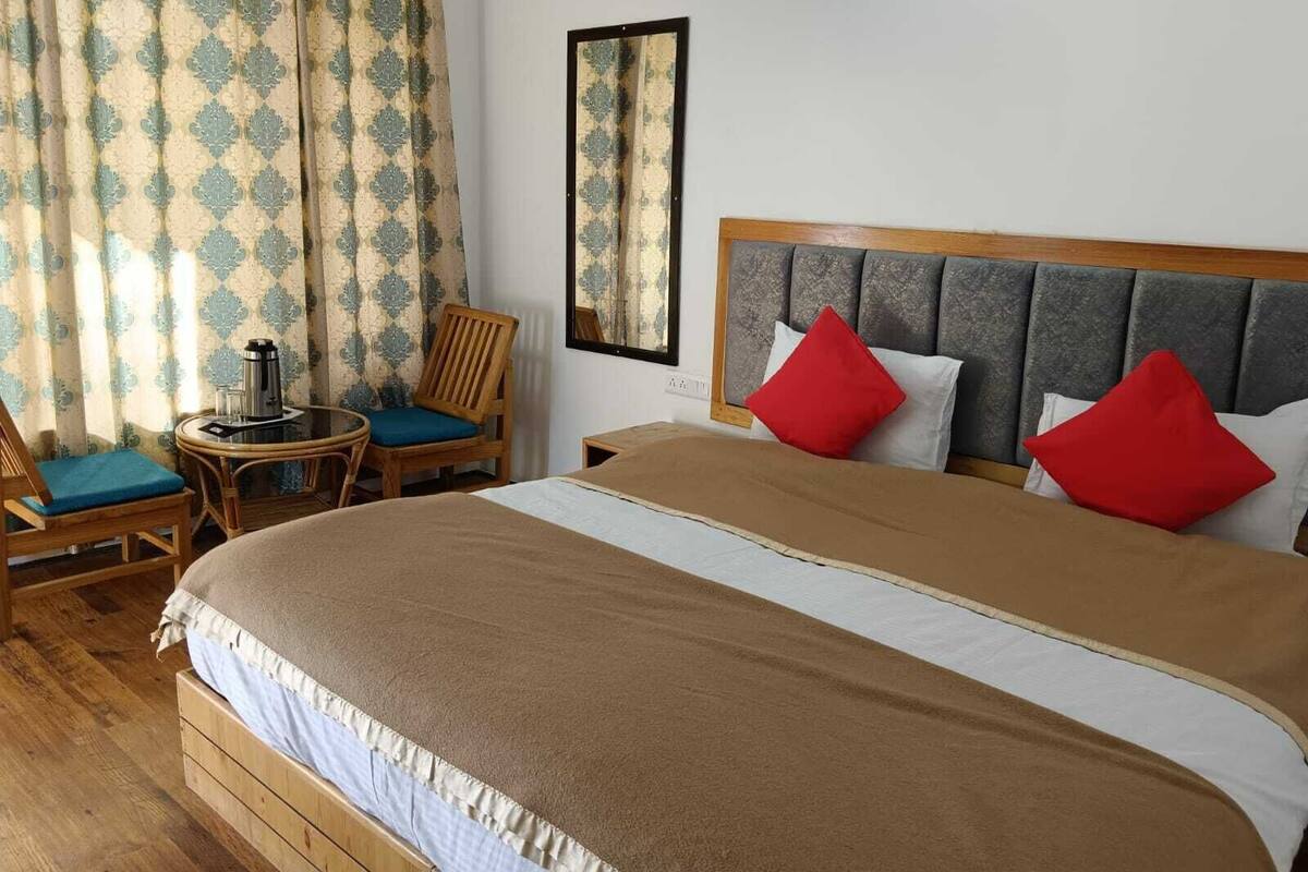 A hotel room with a large bed, two chairs, a small table with a coffee pot, and a mirror on the wall.