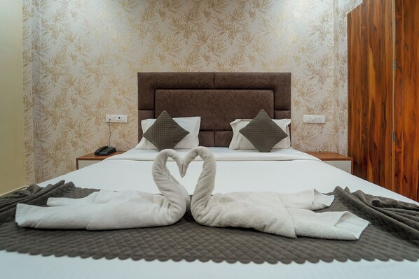 Una cama bien hecha con un cabecero marrón, dos almohadas y un par de toallas dispuestas en forma de corazón sobre un edredón gris.