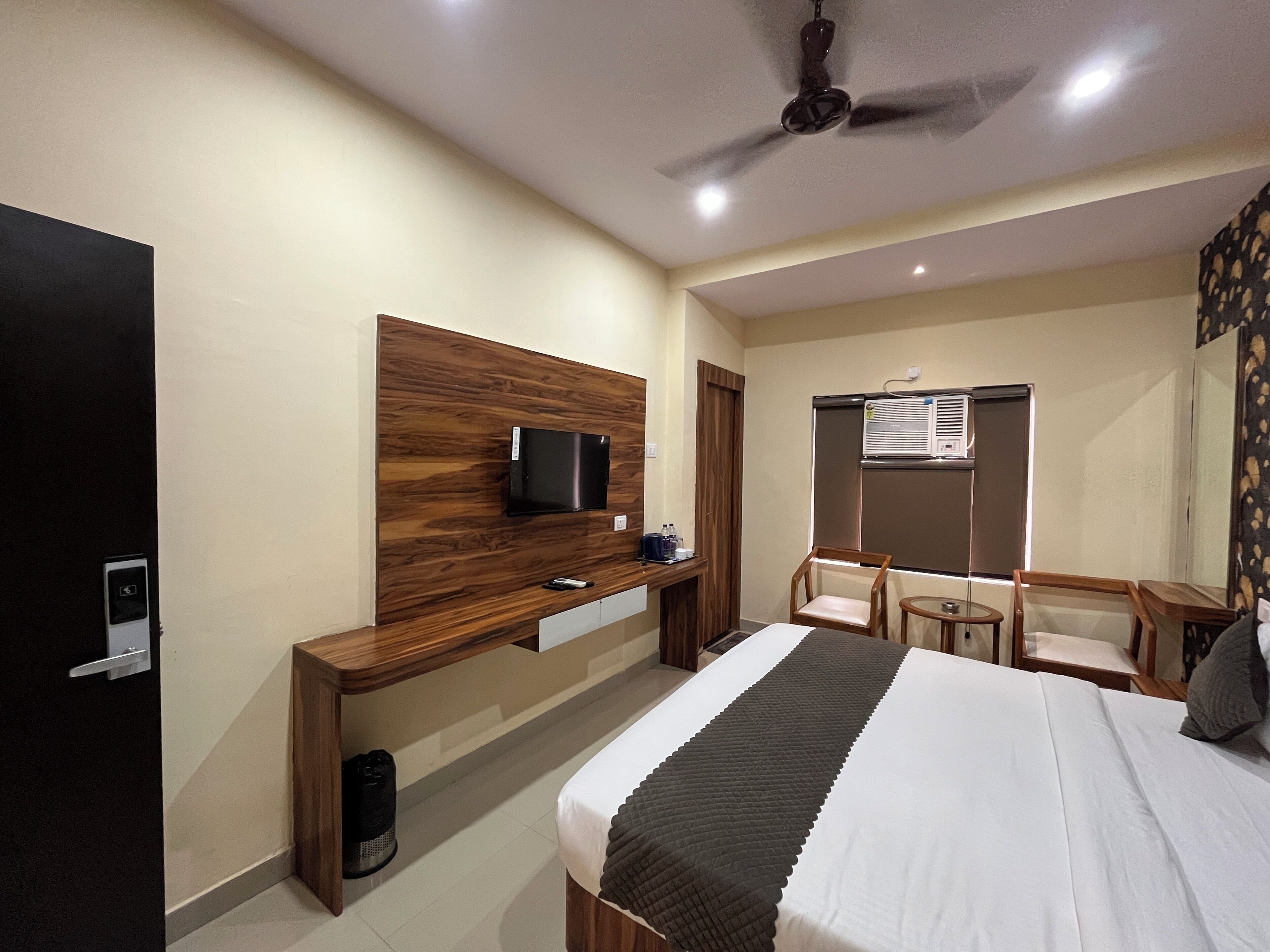 Habitación de hotel con cama, televisión montada en un panel de madera, escritorio con silla y ventilador de techo.