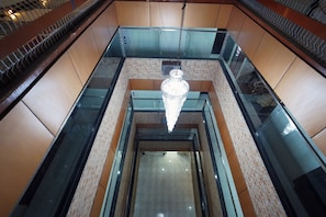 Vista interna di un moderno vano ascensore con cabina in vetro, una lampada a sospensione e un motivo a parete in mattoni.