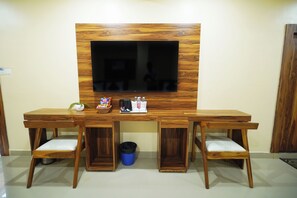 A room with a TV mounted on a wooden wall, a wooden desk with two chairs, and a trash bin. - HOTEL ROSHAN (Varanasi)