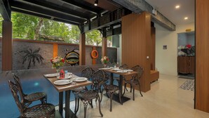Zona pranzo con un lungo tavolo apparecchiato per sei persone, circondato da sedie elaborate, una zona cucina sullo sfondo e vista sugli alberi attraverso ampie finestre.