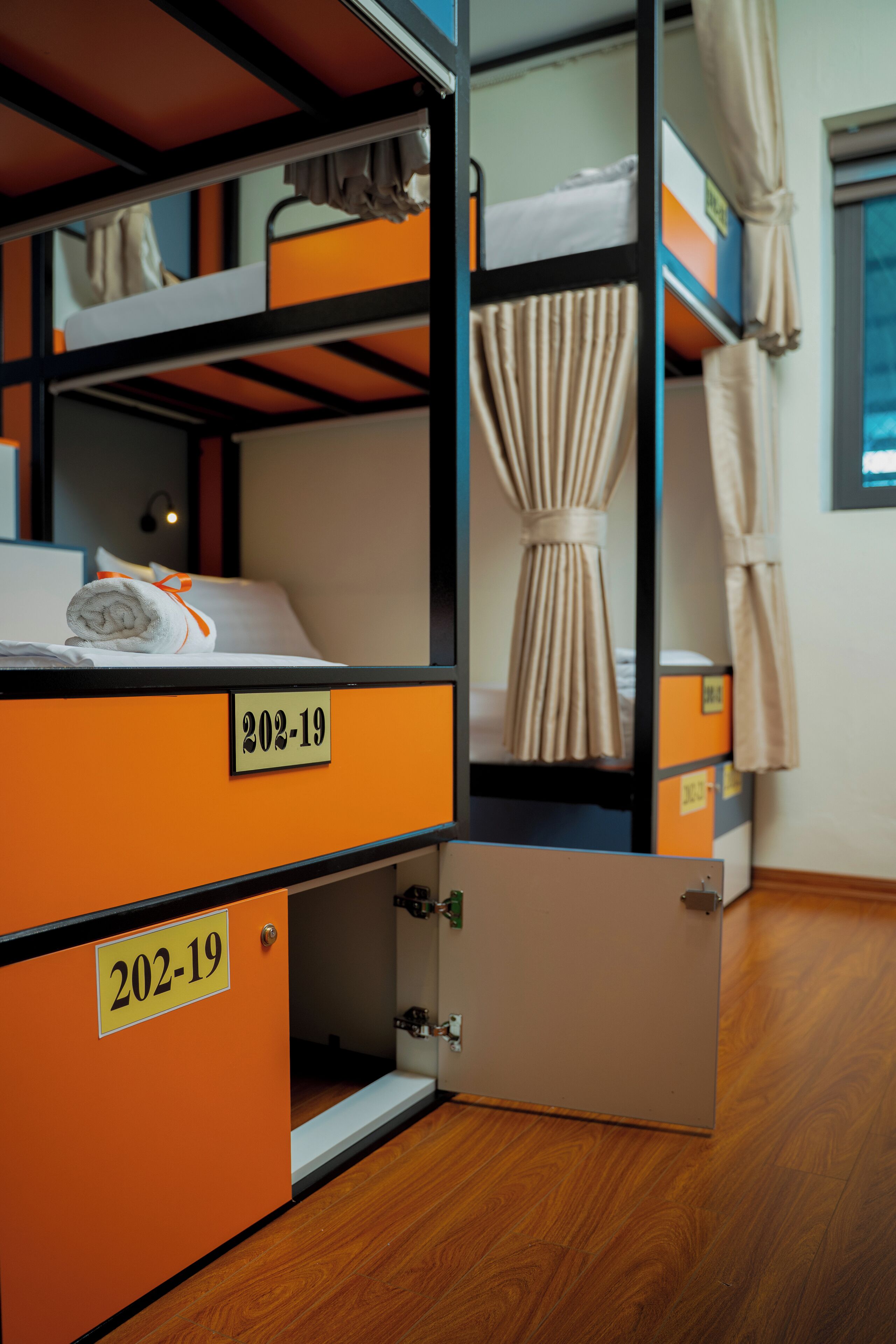 A bunk bed room with a number sign "202-19" on the lower bunk, a curtain on the upper bunk, and a wooden floor.