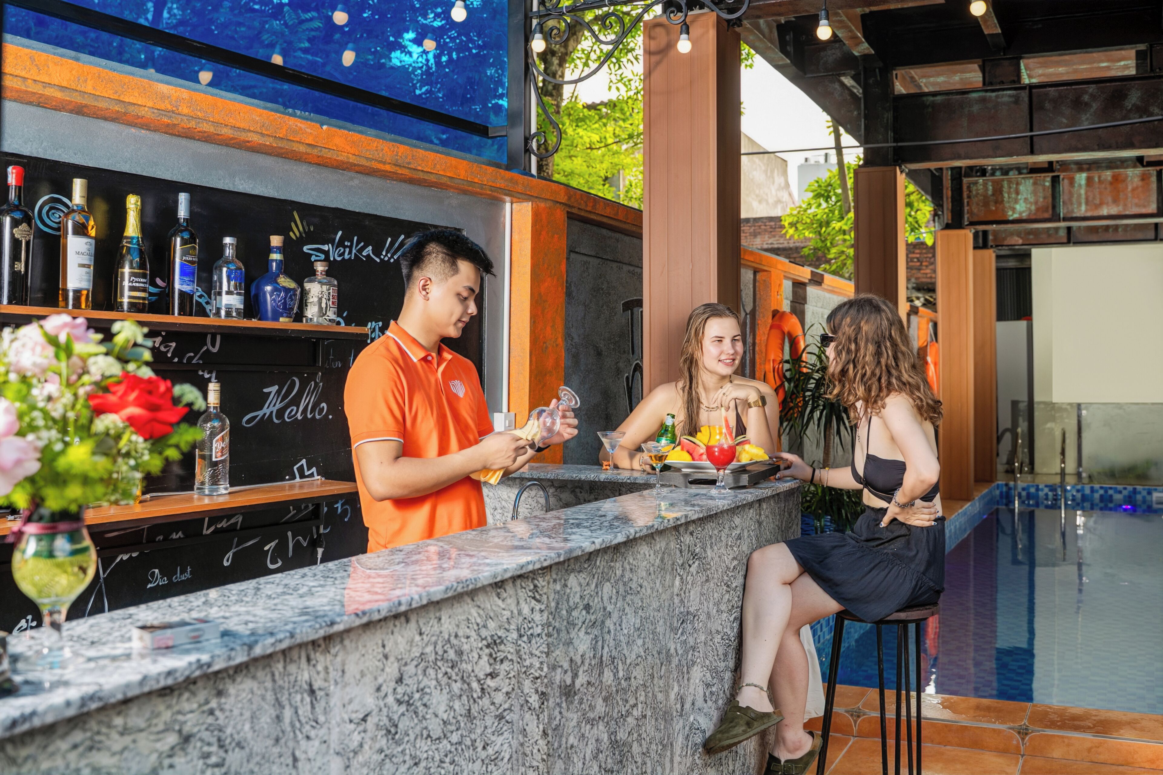En bardisk med en marmorskiva, en bartender i en orange skjorta och en kvinna i en svart klänning som sitter på en barstol.