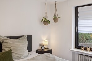Une chambre à coucher avec un lit, des oreillers, une table de chevet avec une lampe et une horloge, deux paniers suspendus contenant des plantes, et une fenêtre avec des stores.