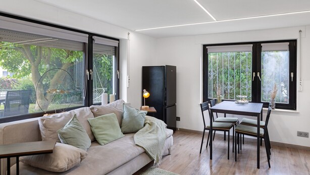 Una sala de estar moderna con sofá, mesa de comedor, sillas, televisor y ventanas grandes.