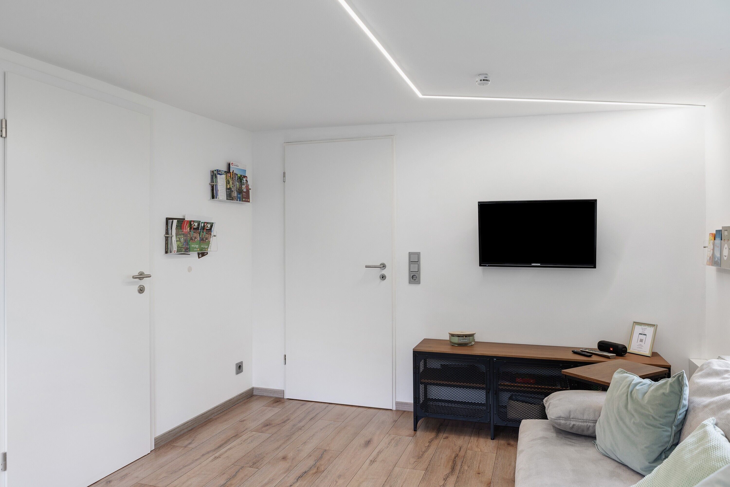 Ett modernt rum med en platt-TV, ett skrivbord i trä, en bekväm soffa och en vit dörr.