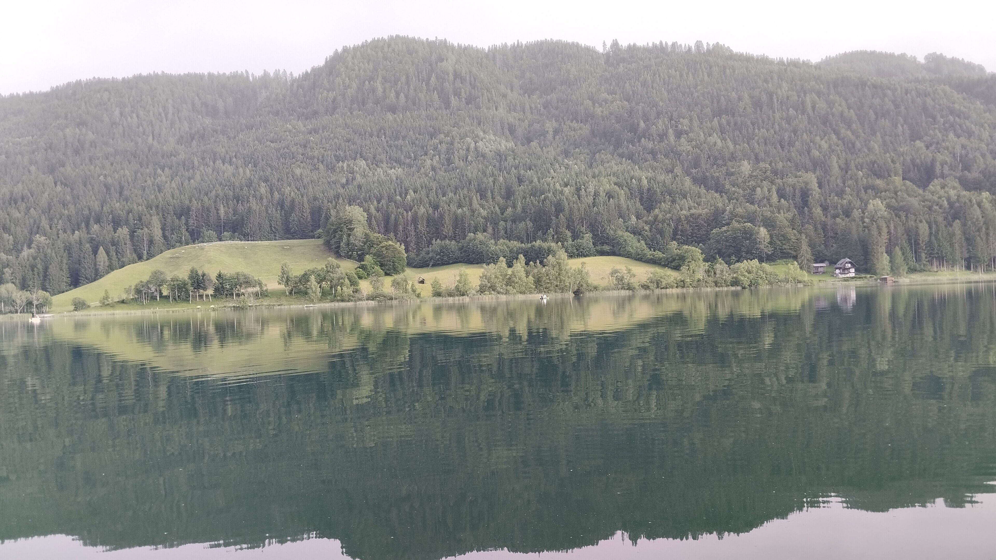 Ein ruhiger See mit einem bewaldeten Hügel im Hintergrund und einem kleinen Haus am Ufer.