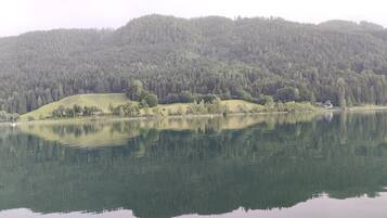 Ein ruhiger See mit einem bewaldeten Hügel im Hintergrund und einem kleinen Haus am Ufer.