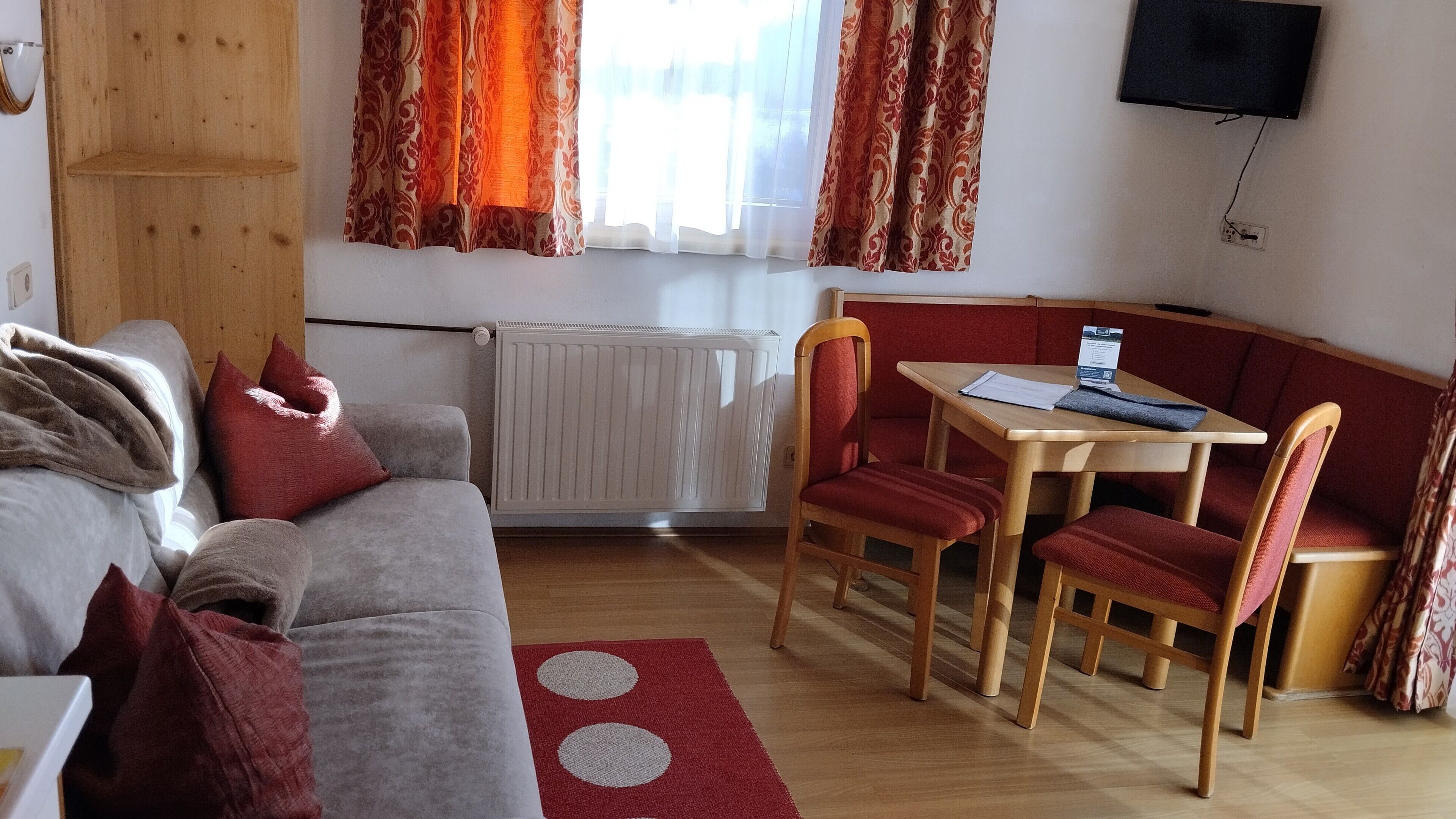 A cozy living room with a sofa, red cushions, a red and white rug, a dining table with chairs, and a radiator.
