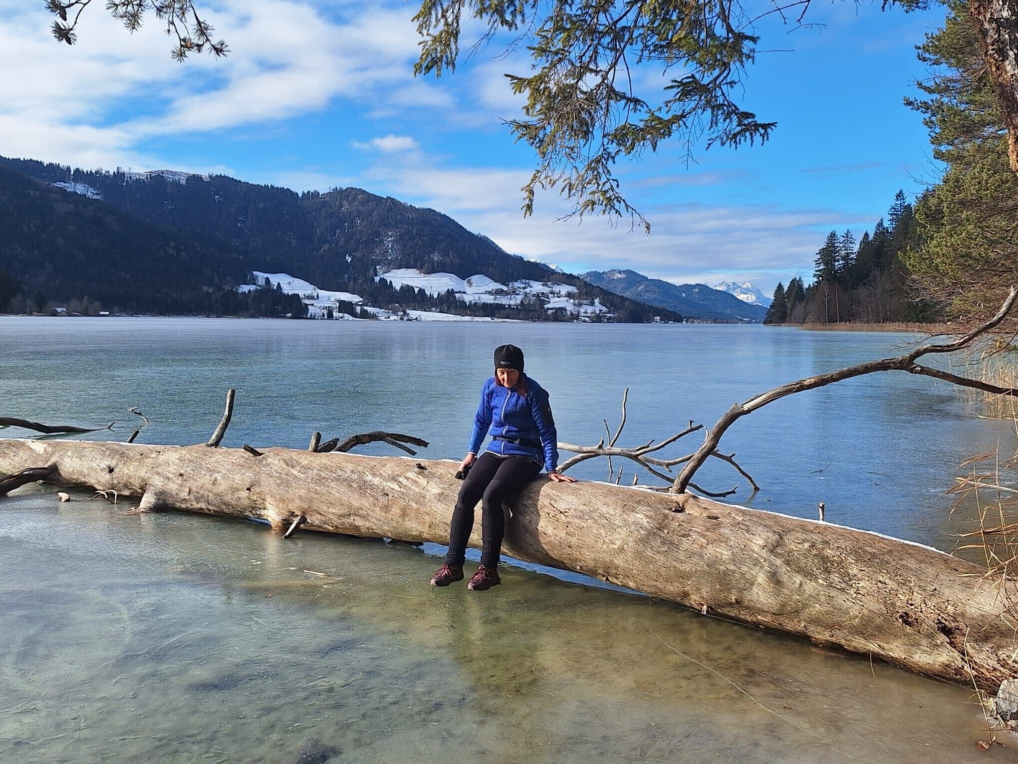 A person sitting on a log by a clear lake with mountains in the background.