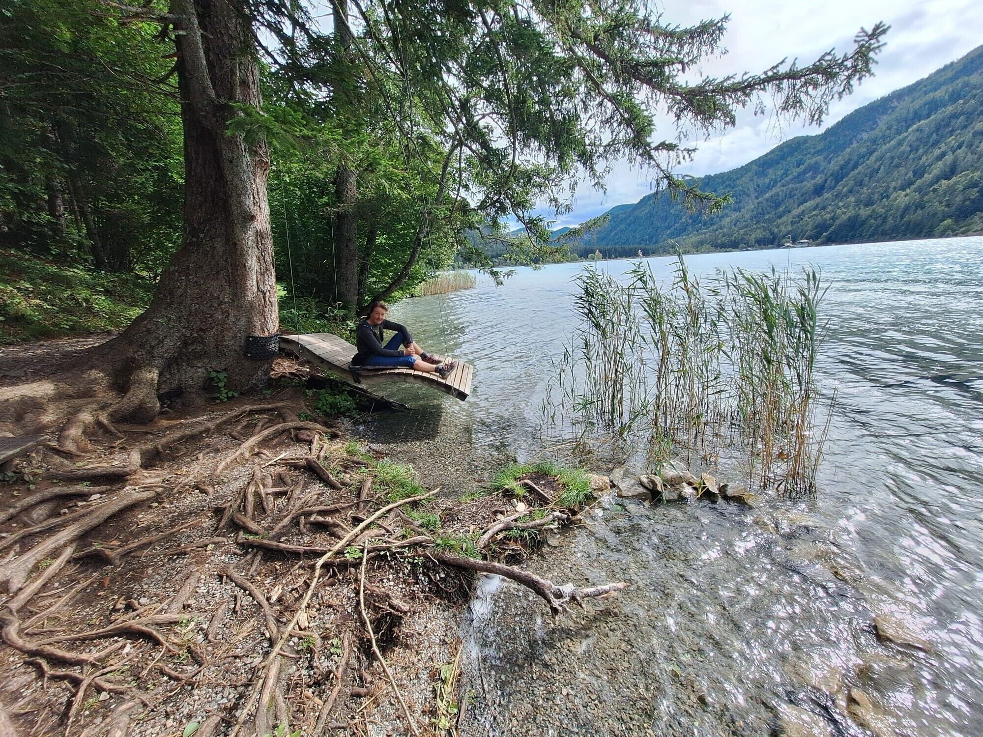 Eine Person sitzt auf einer Holzplattform am Seeufer, umgeben von Bäumen und klarem Wasser.