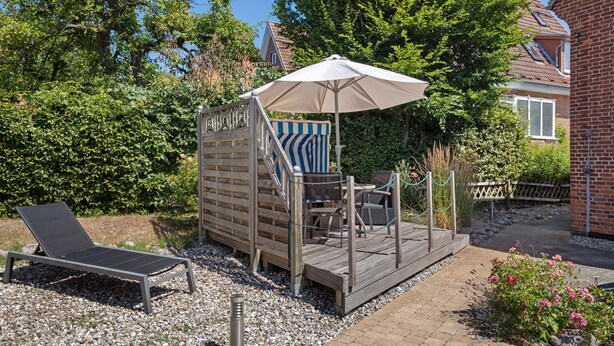 A wooden deck with a lounge chair and a table, surrounded by greenery and a brick building. - Apartment 'Steilküste' with Private Terrace, Private Garden and Wi-Fi (Lübeck Travemünde)