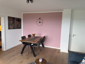 A dining area with a wooden table, six chairs, a vase with flowers, a wall clock, and a map artwork.