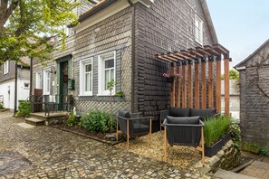 Un patio empedrado con una pérgola de madera, mobiliario exterior y una casa de fachada de ladrillo oscuro y molduras blancas.