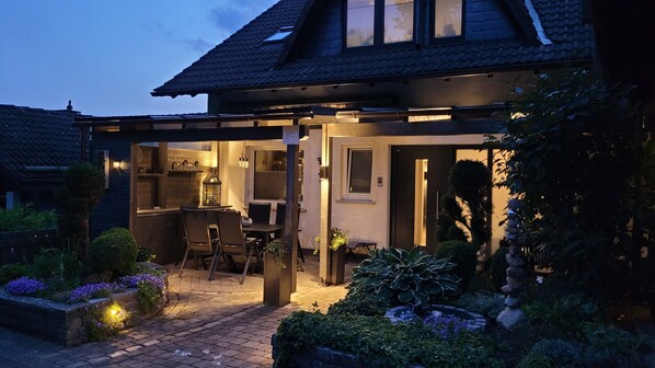 Une maison dotée d’un espace repas extérieur abrité, d’une cuisine ouverte et d’un jardin agrémenté de plantes et d’un élément décoratif en pierre.