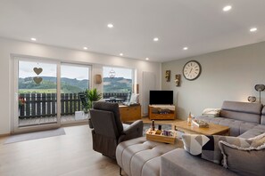 Ein modernes Wohnzimmer mit einer Couch, einem Couchtisch und einem Fernseher. Eine Glastür führt auf einen Balkon mit Blick auf die Landschaft.