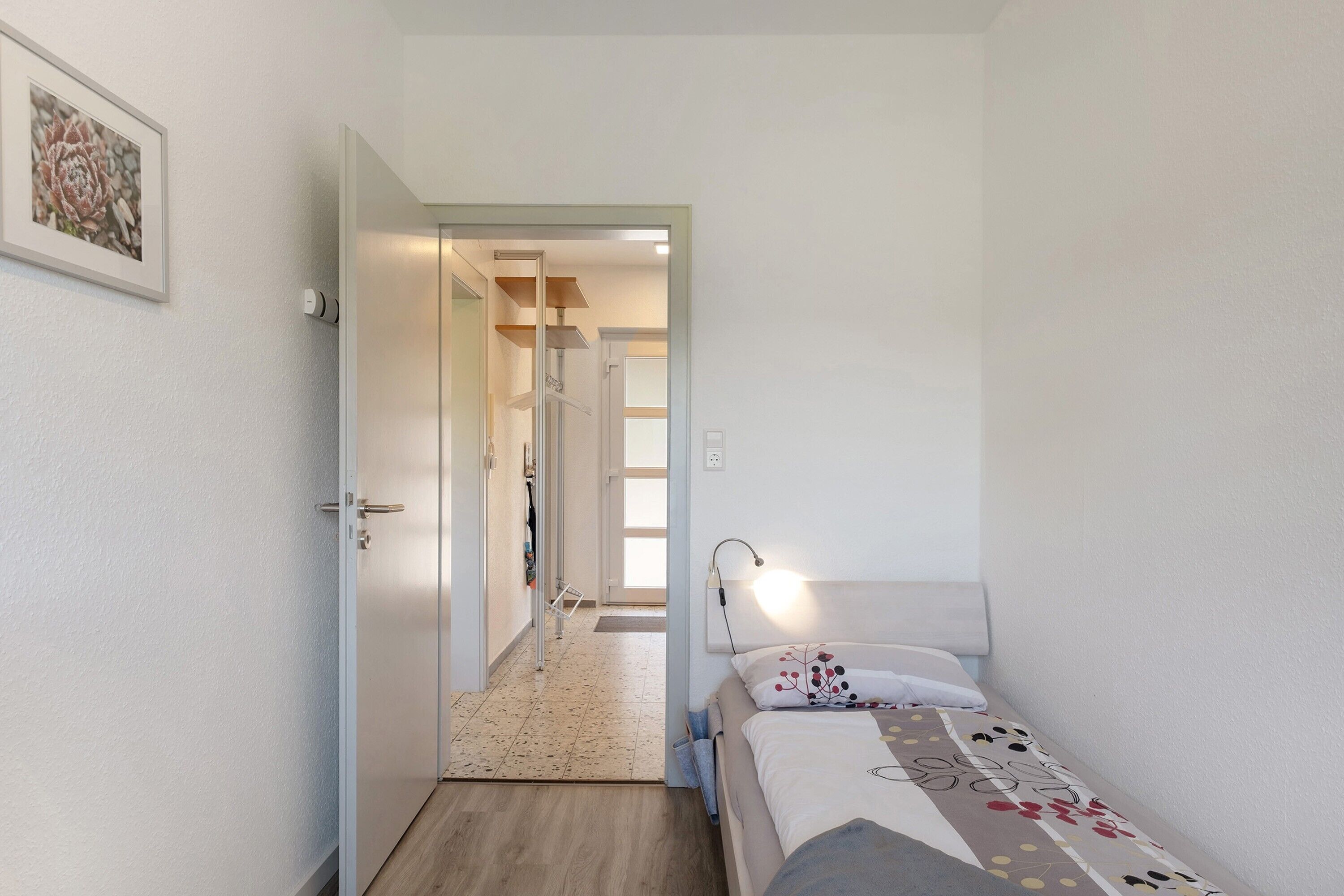 Een slaapkamer met een bed, een bedlamp, een deur naar een gang en een lijst met een afbeelding aan de muur.