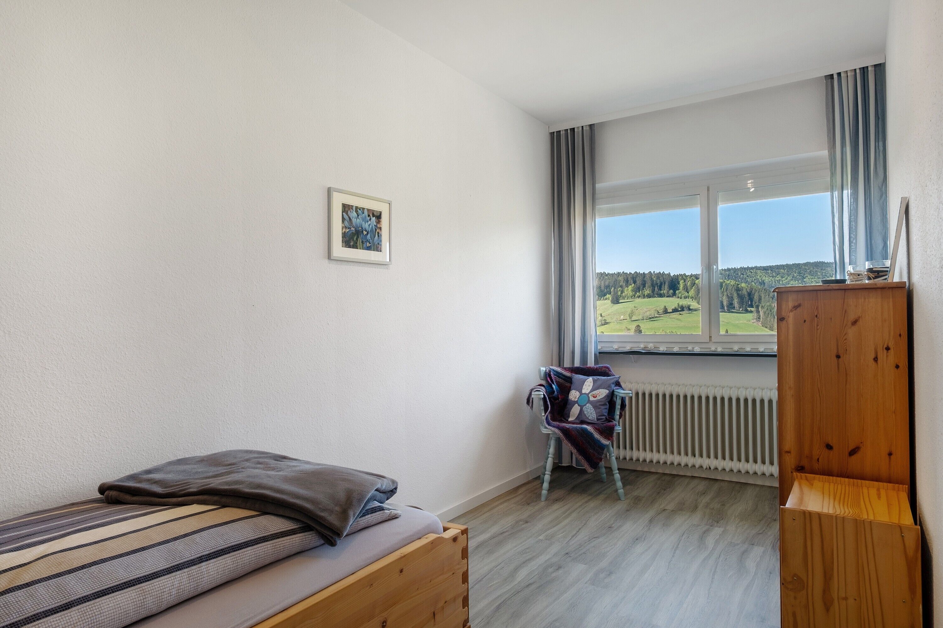A room with a bed, a chair, a radiator, a window with a view of a green landscape, and a wooden cabinet.