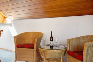 A wicker chair with a red cushion, a glass table with a bottle and two glasses, and a wooden ceiling.