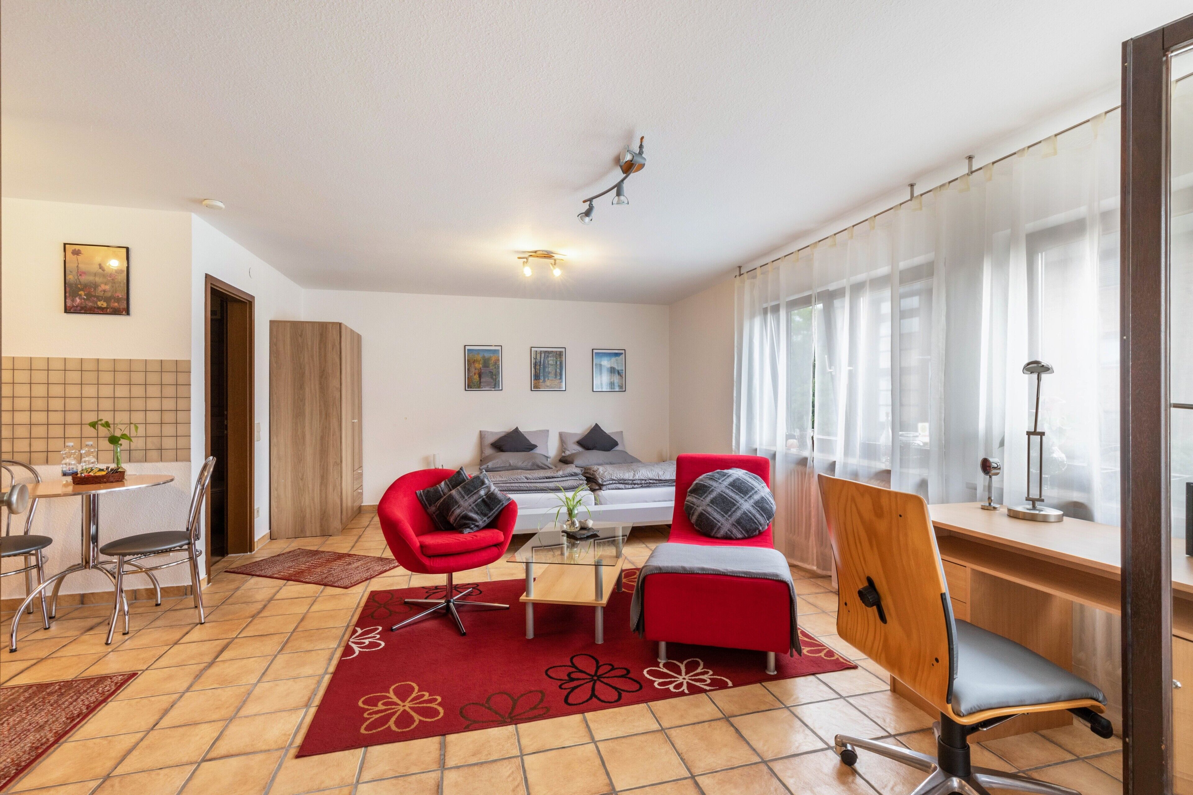 Un saló modern amb un sofà, una butaca i un escriptori. Hi ha una zona de menjador amb caires i una taula. L'habitació té un gran finestral amb cortines.