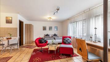 Un saló modern amb un sofà, una butaca i un escriptori. Hi ha una zona de menjador amb caires i una taula. L'habitació té un gran finestral amb cortines.
