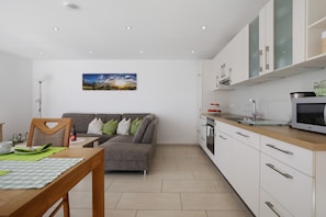 Eine moderne Küche mit weißen Schränken, einem Esstisch mit grünen Platzsets, einem grauen Sofa mit Kissen und einem Gemälde an der Wand.