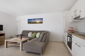 Ein modernes Wohnzimmer mit grauem Sofa, einem Holztisch und einer Küchenzeile mit weißen Schränken und einer Edelstahlspüle.