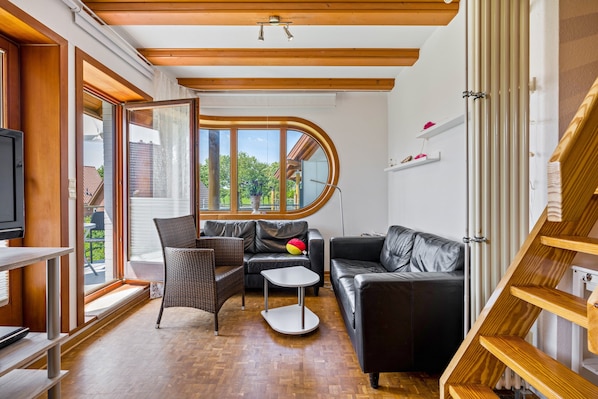 Un salon moderne avec un canapé, un fauteuil et une petite table, un téléviseur et une grande fenêtre donnant sur un paysage verdoyant.