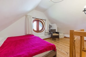 Une chambre avec un plafond en pente, un lit recouvert d’une couverture rouge, une fenêtre avec des rideaux, une chaise et une petite table.
