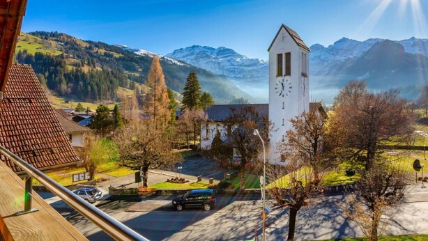A mountain village with a prominent clock tower, a clear view of the surrounding mountains, and a small parking area with cars. - Lovely apartment for 4 guests with WIFI and balcony (Lenk)