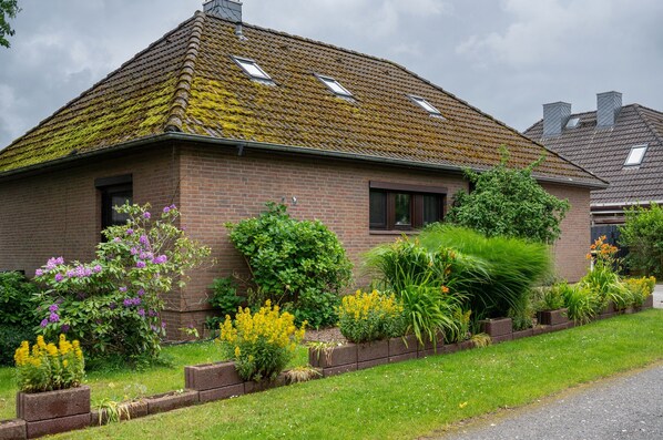 Une maison en briques avec un toit en pente, un jardin où poussent différentes espèces de plantes et une allée pavée.