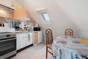 Une cuisine compacte équipée d’une cuisinière, d’un four et de divers ustensiles. On y trouve une table à manger recouverte d’une nappe à motifs de journaux et deux chaises. Une lucarne est visible au plafond.