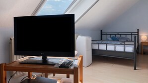 Un dormitorio con cama, un escritorio con monitor de ordenador y ventana con vista al cielo.
